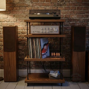 May include: A wooden audio rack with a turntable, amplifier, and vinyl records. The rack has four shelves and black metal supports. Two wooden speakers flank the rack. A digital clock displays 19:05. The background is a brick wall.