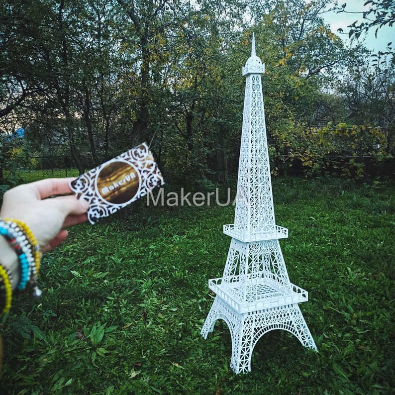 May include: White Eiffel Tower model made of paper or cardboard. The model is standing on a green lawn. A business card with the text "MakerUA" is in the foreground.