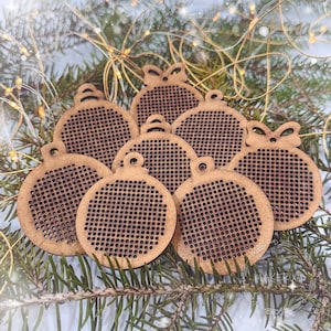 May include: A selection of wooden Christmas bauble cutouts. Each bauble is a light brown colour, featuring a perforated pattern and a small loop for hanging. The baubles are arranged on a bed of green pine needles, evoking a festive atmosphere.