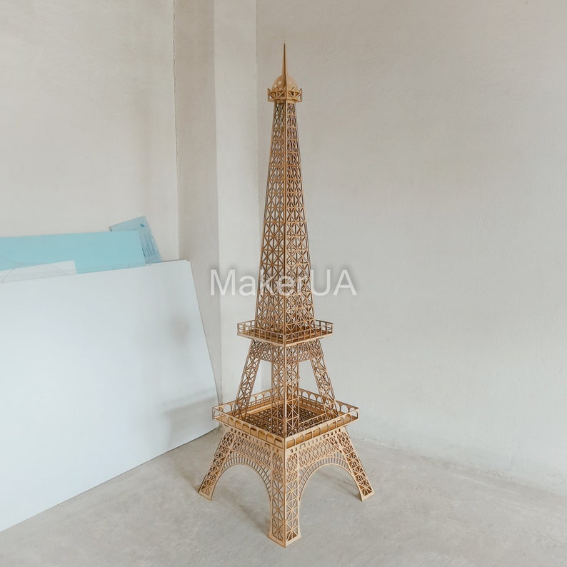 May include: A wooden model of the Eiffel Tower, a famous landmark in Paris, France. The model is made of laser-cut wood and is assembled with intricate details. The model is a light brown color and stands on a white surface.