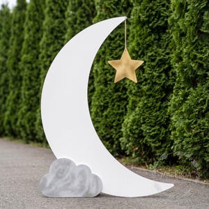 Op de afbeelding: Een grote witte halve maan met een grijze wolk aan de basis en een gouden ster die aan een touwtje hangt. De achtergrond is een rij groene bomen. De maan en ster zijn waarschijnlijk decoratieve elementen voor een kinderkamer of evenement.