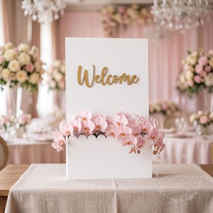 May include: A rectangular white sign featuring the word Welcome in gold script lettering. The sign has a scalloped top edge on the lower section, which is adorned with a cluster of light pink orchid flowers arranged across the front surface.