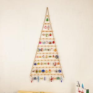 May include: A wooden Christmas tree-shaped ornament display with multiple small, colorful ornaments hanging from it. The display is made of wooden dowels and twine, creating a rustic aesthetic. The ornaments are various shapes and colors.