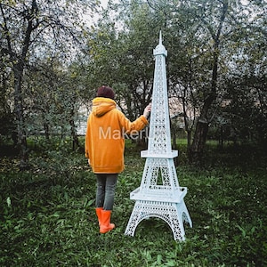 May include: A white paper model of the Eiffel Tower stands in a grassy area. A person wearing a yellow jacket and orange boots stands behind the model, looking at it.