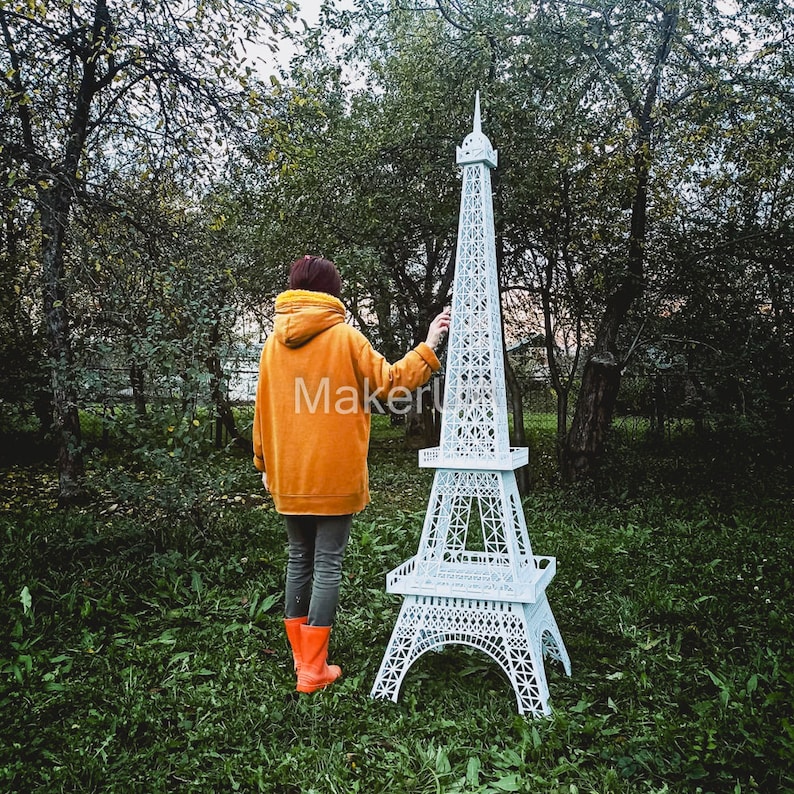 May include: A white paper model of the Eiffel Tower stands in a grassy area. A person wearing a yellow jacket and orange boots stands behind the model, looking at it.