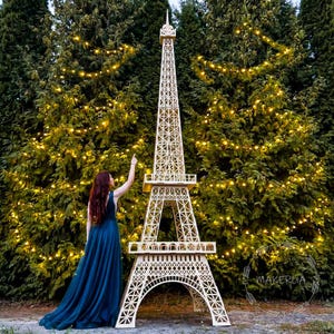 Può includere: Una grande replica in legno della Torre Eiffel, dal design intricato, si erge su uno sfondo di lussureggianti alberi verdi adornati da lucine scintillanti. La torre è di colore beige chiaro, con dettagliati lavori a traliccio.