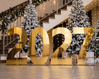 May include: Large, freestanding gold-colored reflective numbers spelling 2027. Each digit has a smooth, metallic finish and stands on a flat base. The text MAKERUA is visible in the bottom right corner within a circular decorative border.