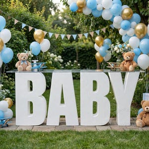 Op de afbeelding: Een wit 'BABY'-bord met een doorzichtige bovenkant, versierd met blauwe, gouden en witte ballonnen, teddyberen en speelgoedvliegtuigen. Een feestelijke banner hangt erboven en creëert een feestelijke sfeer voor een babyshower of gender reveal party.