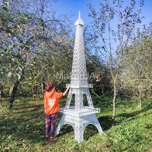 May include: A white, metal Eiffel Tower replica standing in a grassy area. A person is standing near the tower, looking at it.