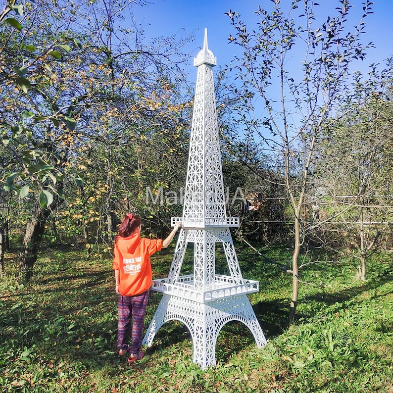 May include: A white, metal Eiffel Tower replica standing in a grassy area. A person is standing near the tower, looking at it.