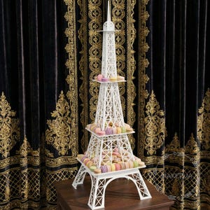 May include: A white Eiffel Tower-shaped macaron stand, filled with colourful macarons. The stand has three tiers and is made of a white material. The macarons are in pastel colours, including pink, yellow, and green. The stand is on a wooden surface.