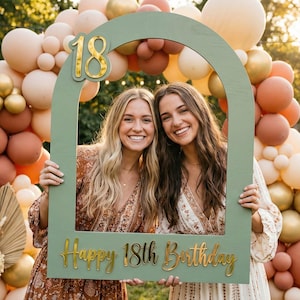 Może przedstawiać: Zielona drewniana ramka na zdjęcia ze złotymi literami z napisem "18" i "Happy 18th Birthday". Dwie uśmiechnięte kobiety trzymają ramkę przed łukiem z balonów w odcieniach brzoskwini, złota i bieli.
