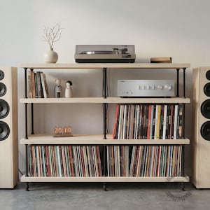 May include: A light wood and black metal record storage unit with a turntable, amplifier, and speakers. The shelves are filled with vinyl records, books, and decorative items. The unit has a minimalist design and is suitable for a home entertainment setup.