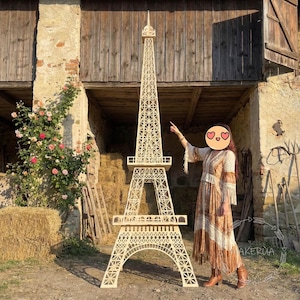 May include: A large, intricately designed wooden Eiffel Tower replica. The model is light-colored, with detailed cutouts mimicking the original structure. It stands tall against a rustic backdrop, suggesting a decorative or souvenir item.