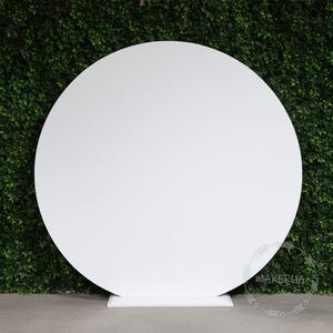 May include: A large, white, circular backdrop stands on a white base, set against a backdrop of green foliage. The backdrop is divided into two vertical panels. The word "MAKERUA" is printed on the lower right corner.