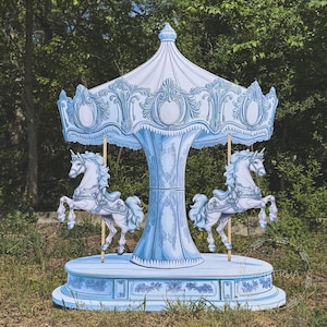 May include: A light blue carousel with white accents and two unicorn figures. The carousel has a decorative canopy and base, with intricate detailing. The carousel is set outdoors, surrounded by greenery.