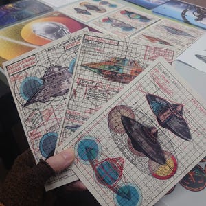 May include: Three rectangular art prints on grid paper featuring technical illustrations of flying saucers. The prints include hand-drawn sketches, red and blue geometric overlays, and handwritten text labels such as Bell-Shaped Flying Saucer, Element 115, and Acorn + Torus Flying Saucer.