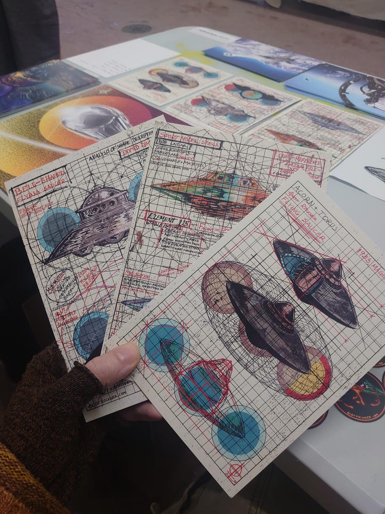 May include: Three rectangular art prints on grid paper featuring technical illustrations of flying saucers. The prints include hand-drawn sketches, red and blue geometric overlays, and handwritten text labels such as Bell-Shaped Flying Saucer, Element 115, and Acorn + Torus Flying Saucer.