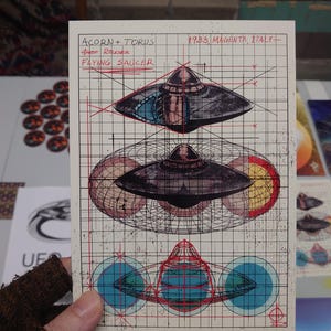 May include: A rectangular print on grid paper featuring technical illustrations of a flying saucer. Text reads ACORN + TORUS, 1933, MAGENTA, ITALY, and FLYING SAUCER. The design includes three stacked views of the craft with red and blue geometric overlays.