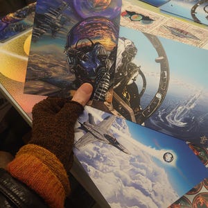 May include: A set of three colourful prints featuring a pilot in a cockpit, a fighter jet flying through clouds, and a geometric design with a galaxy background.