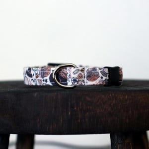 Puede incluir: Un collar para perro estampado con una anilla en D de metal y una hebilla de pl&aacute;stico negro. El collar presenta un fondo blanco con formas marrones y grises. Descansa sobre un peque&ntilde;o taburete de madera oscura.