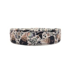 Puede incluir: Un collar para perros beige con un patr&oacute;n repetido de ilustraciones con tem&aacute;tica de brujas. El dise&ntilde;o incluye calderos, botellas de pociones, bolas de cristal, sombreros de bruja y estrellas. El collar est&aacute; hecho de una tela resistente.