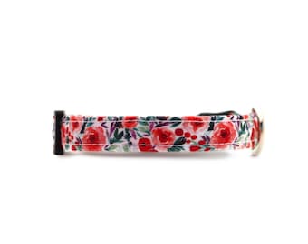 May include: A floral pet collar featuring a white background with red roses, green leaves, and red berries. The collar has a black plastic buckle and a silver metal D-ring. The collar is designed for a small dog or cat.