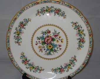 May include: A white porcelain plate with a gold rim and a floral design. The center of the plate features a large floral motif with pink, yellow, and blue flowers. The plate is surrounded by a smaller floral border.
