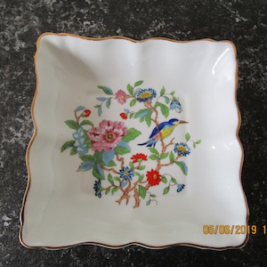 Op de afbeelding: Een wit vierkant schaaltje met een gouden rand en een bloemmotief met een blauwe vogel, roze, rode en blauwe bloemen en groene bladeren.