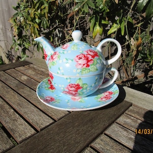 May include: A light blue tea set with a floral rose pattern. The set includes a teapot, teacup, and saucer. The teapot has a white handle and a matching lid. The teacup sits inside the teapot, and the saucer is beneath.