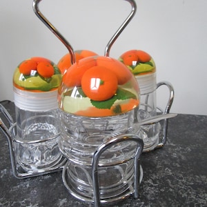 May include: A set of four clear glass condiment jars with orange and green fruit designs on the lids. The jars are held together by a chrome metal stand with a handle.