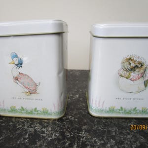 May include: Two white rectangular metal storage tins featuring illustrations of Jemima Puddle-Duck and Mrs. Tiggy-Winkle. Each tin has a gold-toned rim, a lid, and a base border of green grass and small flowers. The names are printed below each character.