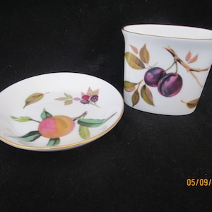 Puede incluir: Un juego de dos platos de cerámica blanca con ribete dorado. El plato más pequeño es de forma ovalada y presenta un diseño pintado de ciruelas en una rama. El plato más grande es redondo y presenta un diseño pintado de un melocotón en una rama.