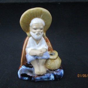May include: Ceramic figurine of a man wearing a straw hat and sitting with a basket. The figurine is painted in shades of brown, blue, and white.