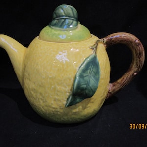 Op de afbeelding: Een citroenvormige keramische theepot met een getextureerd gele oppervlak en een groene deksel. De handgreep is ontworpen als een tak, en een groen blad is bevestigd. Een decoratief keukenartikel.