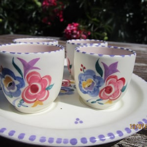 Puede incluir: Un juego de cuatro tazas de cerámica blanca con diseños florales en rosa, azul y morado, sobre una bandeja a juego. La bandeja y las tazas tienen un borde lavanda. La fecha 12/07/2025 es visible en la bandeja.
