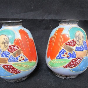 May include: Two small, round, ceramic vases with a blue glaze. Each vase features a hand-painted design of a man in a colorful robe with gold accents. The vases have a black rim.