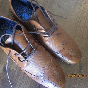 May include: A pair of brown leather lace-up shoes with a brogue design. The shoes have a blue and white patterned insole with the word "Taylor" printed on it.