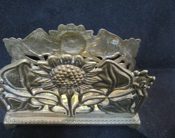 Puede incluir: Un porta servilletas de metal dorado con un diseño de girasol. El porta servilletas tiene un diseño decorativo calado con un girasol en el centro y hojas alrededor.