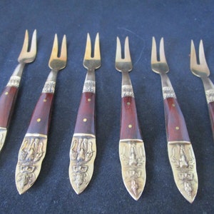 May include: Set of seven small brass forks with wooden handles. The forks feature ornate designs and are ideal for serving appetisers or desserts.