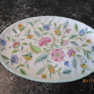 May include: An oval serving platter with a white base, decorated with a floral pattern in pink, blue, yellow, and purple. Green leaves and stems complement the flowers. The rim is a pale green. The date 01/11/2025 is printed on the bottom right.