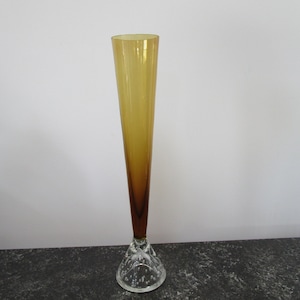 Op de afbeelding: Een hoge, amberkleurige glazen vaas met een heldere glazen voet. De voet heeft een textuur met bubbels.