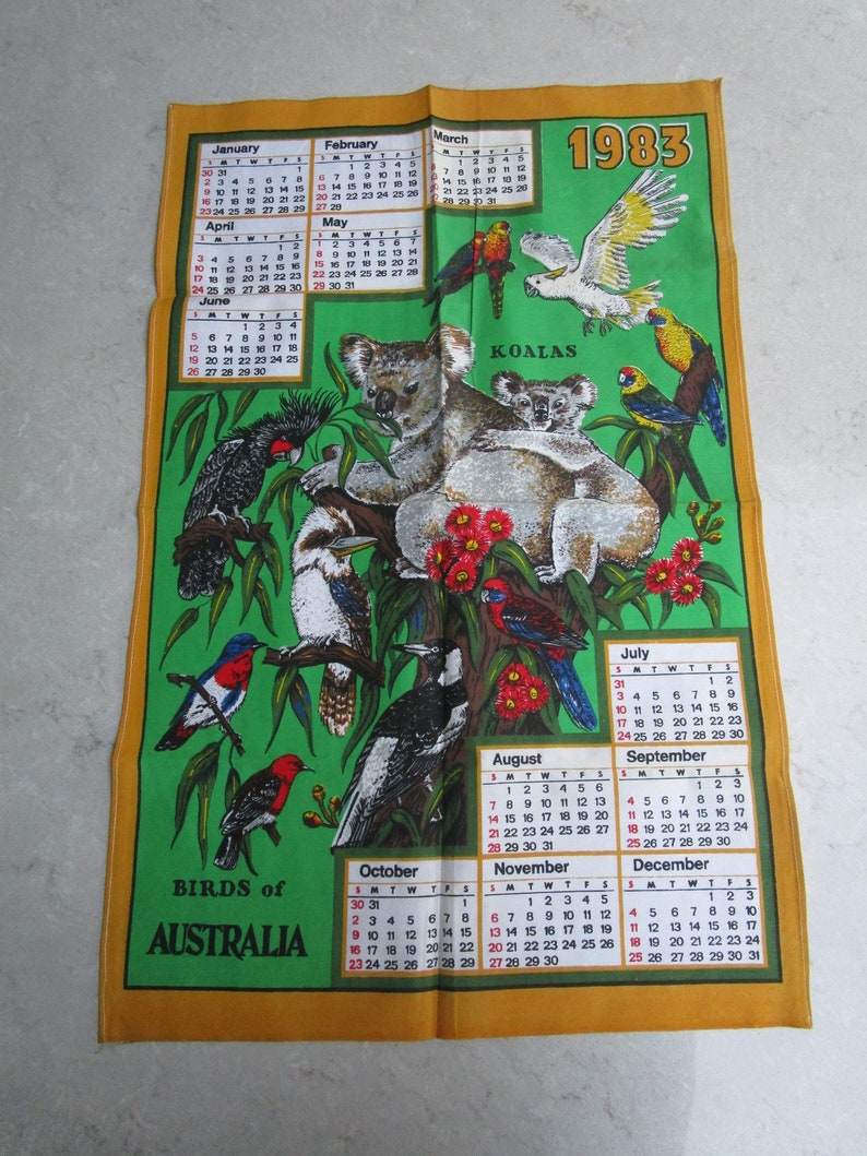 May include: A vintage 1983 calendar tea towel featuring a green background with illustrations of Australian birds and koalas. The calendar includes the months of January through December with the year 1983 printed in large, bold letters at the top.
