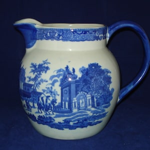 Puede incluir: Una jarra de cerámica azul y blanca con asa. La jarra tiene un diseño floral y una escena de un edificio con árboles y personas.