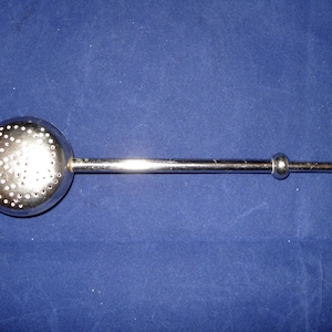 Könnte beinhalten: Ein silberner Tee-Ei mit einem runden, perforierten Sieb und einem langen Griff mit zwei runden Knöpfen am Ende.