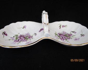 May include: White porcelain serving dish with a handle, featuring a divided design and scalloped edges. The dish is decorated with clusters of purple violets and green leaves, and trimmed with gold accents.