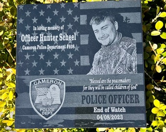 May include: A black granite memorial plaque with a photo of a man in camouflage, the text "In loving memory of Officer Hunter Scheel Cameron Police Department #608", a police badge, and the quote "Blessed are the peacemakers for they will be called children of God." The plaque also includes the text "Police Officer End of Watch 04/08/2023".