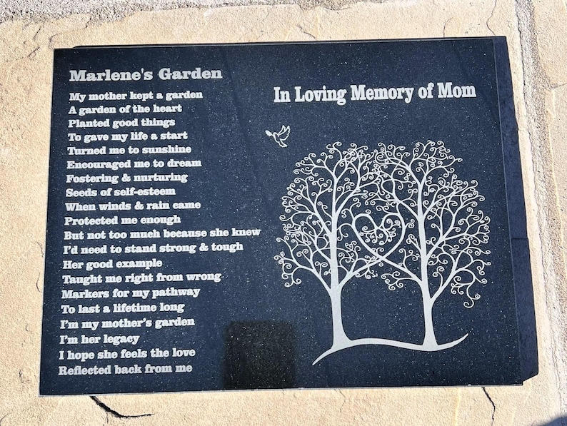 May include: Black granite memorial plaque with a poem about a mother's garden and a white silhouette of two trees with a heart shape in the center. The text reads "Marlene's Garden" and "In Loving Memory of Mom".