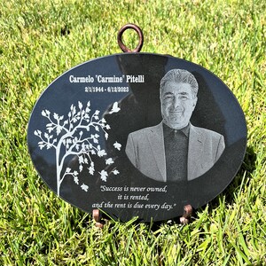 May include: Black oval granite memorial plaque with a photo of a man, a tree with white leaves, and the text "Carmelo "Carmine" Pitelli 2/1/1944-6/12/2023" and "Success is never owned, it is rented, and the rent is due every day."