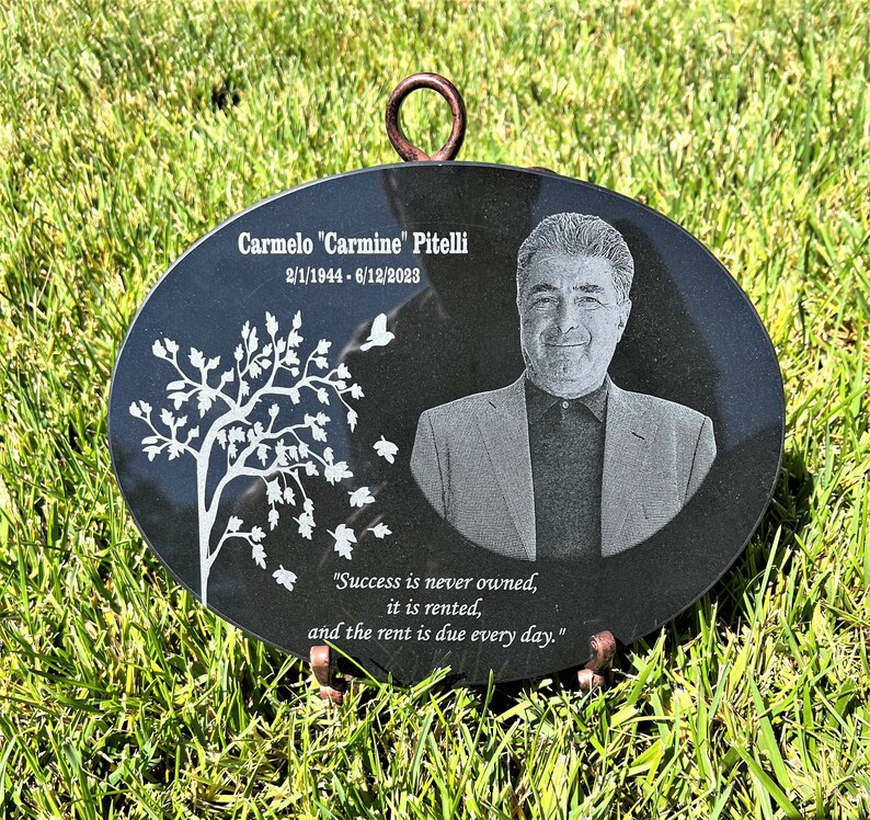May include: Black oval granite memorial plaque with a photo of a man, a tree with white leaves, and the text "Carmelo "Carmine" Pitelli 2/1/1944-6/12/2023" and "Success is never owned, it is rented, and the rent is due every day."